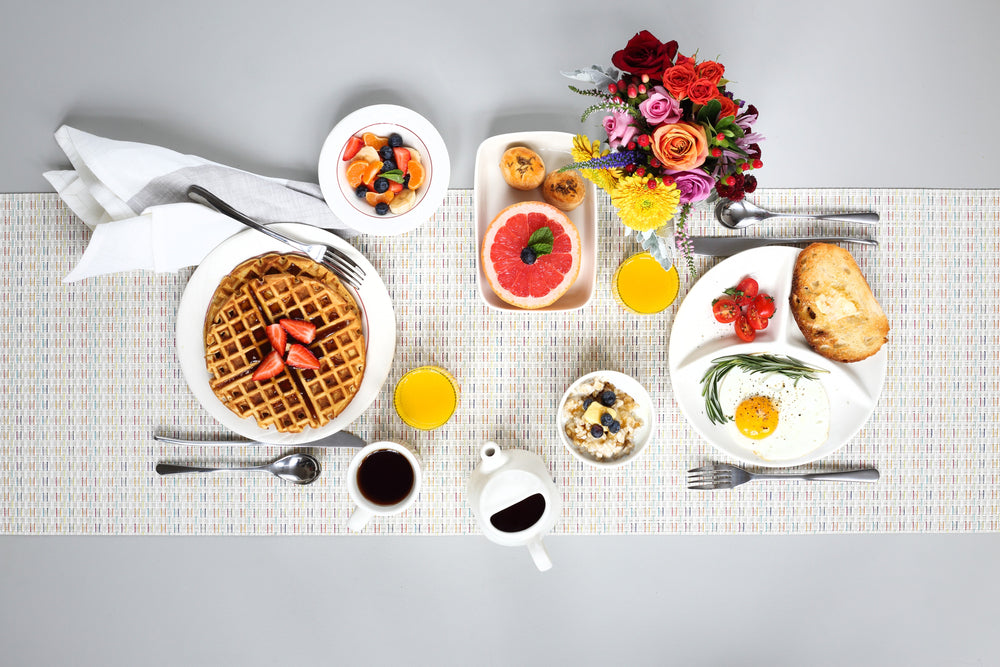 Breakfast table setting with waffles, fruit, coffee, and flowers on a white tablecloth.